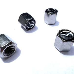 Puede incluir: Cuatro tapones de válvula de neumático plateados con el logotipo de Mazda. Las tapas son hexagonales y tienen un interior roscado. El logotipo de Mazda es una 'M' estilizada dentro de un círculo.