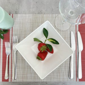 May include: A white square plate with three red strawberries and a green leaf sprig on a beige placemat with red stripes. Two silver forks and two silver knives are on the placemat.