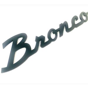 May include: Black, cursive-style "Bronco" emblem. The emblem is a single piece, with a matte finish. The letters are connected, and the design is slightly angled against a white background.