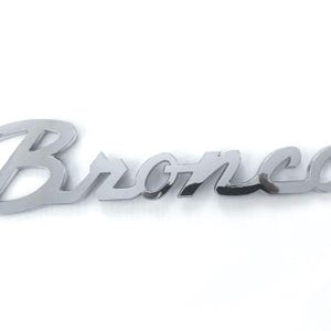 Puede incluir: Emblema cromado "Bronco", un accesorio para automóvil, sobre un fondo blanco. La palabra "Bronco" está escrita en una fuente cursiva estilizada. El emblema es probablemente para un vehículo.