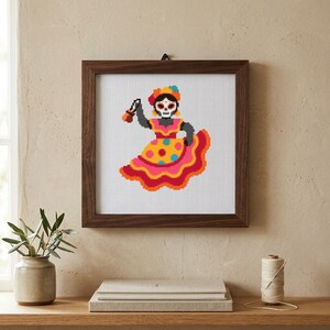 Puede incluir: Un patrón de punto de cruz en un marco de madera oscura que muestra una figura pixelada con cara de calavera, vistiendo un vestido amarillo y rosa vibrante con lunares coloridos y un tocado floral, sosteniendo un objeto marrón.