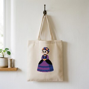 Puede incluir: Un bolso de tela beige con un gráfico de pixel art de una figura con cara de calavera, que lleva un vestido morado y negro con un tocado a juego. La figura tiene el pelo naranja y se muestra en una postura formal y erguida.