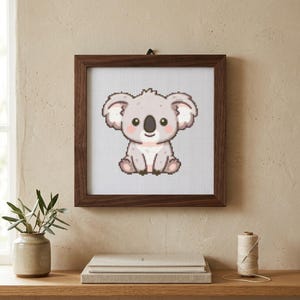 Puede incluir: Un marco de madera cuadrado que contiene un patrón de punto de cruz de un koala gris claro con orejas grandes, nariz negra y mejillas rosadas. El koala está representado en un estilo pixelado sobre un fondo de cuadrícula blanca.