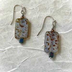 May include: A pair of silver dangle earrings with rectangular glass beads. The beads are translucent and have a brown and white pattern. The beads are accented with a small, dark blue bead at the bottom.