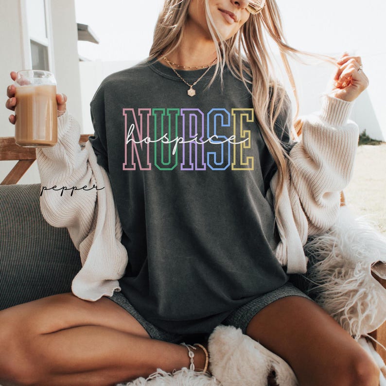 May include: A dark grey t-shirt with the text "NURSE hospice" in rainbow colours. The text is outlined in black and has a vintage collegiate style.