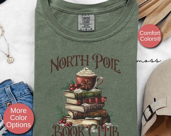 May include: A green t-shirt with the text "North Pole Book Club" in brown lettering. The design features a stack of books with a cup of hot chocolate on top, surrounded by red berries and snow.