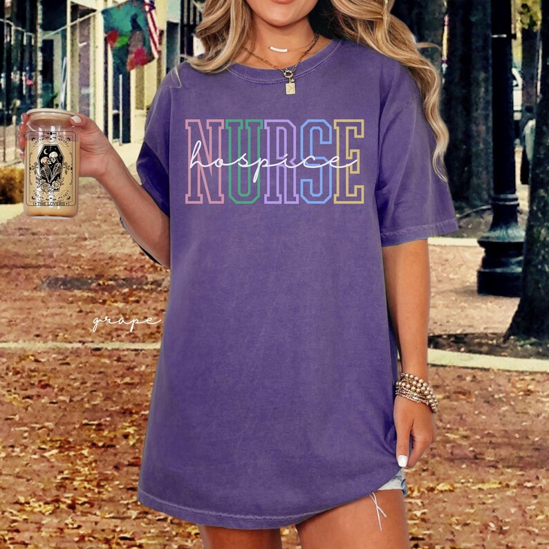 May include: A purple t-shirt with the text "NURSE hospice" in rainbow colours. The shirt is a relaxed fit and has a crew neck.