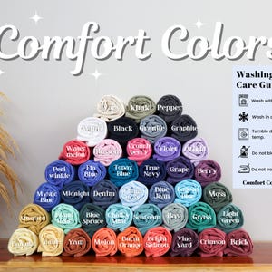 Pode incluir: Uma exibi&ccedil;&atilde;o de camisetas enroladas em v&aacute;rias cores, incluindo preto, branco, azul, verde, vermelho e amarelo. As camisetas est&atilde;o dispostas em forma de pir&acirc;mide e etiquetadas com seus nomes de cores. O texto "Comfort Colors 1717" &eacute; vis&iacute;vel na parte inferior da imagem.