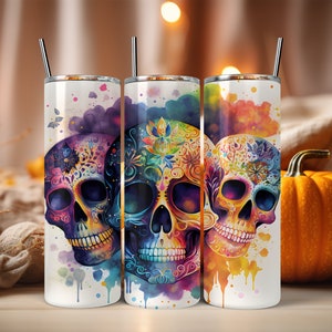 May include: Three stainless steel tumblers with colorful watercolor designs of sugar skulls. The skulls are decorated with intricate floral patterns and have a vibrant, multi-colored appearance.