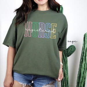 Pode incluir: Camiseta verde-s&aacute;lvia com o texto "NURSE registered" em cores do arco-&iacute;ris.