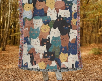 May include: A woven throw blanket featuring a repeating pattern of stylized cats in various colors including black, white, brown, yellow, and blue. The blanket has a textured, tapestry-like appearance with fringed edges on the left and right sides.