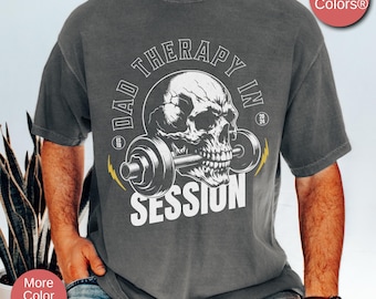 May include: Pepper-colored t-shirt with a graphic of a skull lifting a barbell, with the text "DAD THERAPY IN SESSION". The design includes lightning bolt accents and the Comfort Colors logo.