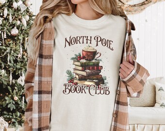 May include: A white t-shirt with a graphic of a stack of books, a cup of coffee, and the text "North Pole Book Club".