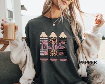 May include: A dark gray t-shirt with a design of three pencils. Each pencil is decorated with a different pattern: black and white stripes, leopard print, and black and white stripes with pink flowers. The text "Mrs. Parker" is printed in pink on the shirt.