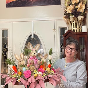 May include: An Easter-themed decorative arrangement with plush bunny figures, colourful eggs, carrots, and pink and green floral accents. The display is set in a wooden tray, creating a festive seasonal display.