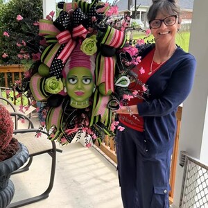 Può includere: Una ghirlanda decorativa di Halloween con al centro la testa di un personaggio dalla pelle verde e capelli rosa, circondata da nastri a righe neri, rosa e verde lime, fiocchi a pois, fiori rosa artificiali e fogliame nero.