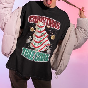 May include: A black t-shirt featuring a white Christmas tree cake design. The text "Christmas Tree Cake" is written in green and red lettering.