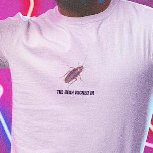 May include: A person wearing a white t-shirt with a cockroach graphic and the text "The Bean Kicked In". The person is wearing green shorts and neon sunglasses. The background is a purple wall with neon pink and blue geometric shapes.