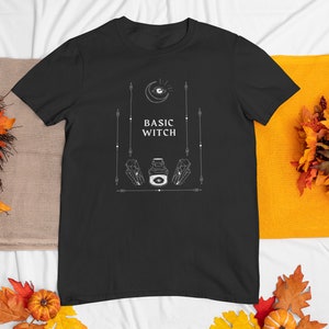 以下が含まれることがあります： 三日月、目、クリスタル、そして「Basic Witch」というテキストが描かれた白いグラフィックデザインが施された黒いTシャツです。