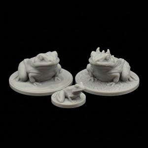 May include: Three gray frog figurines of varying sizes. Two larger frogs sit on lily pads, while a smaller frog rests on a smaller lily pad. The frogs have detailed features and are set against a black background.