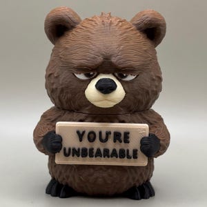 May include: A brown 3D printed bear figurine with a textured fur finish, black paws, and a cream-colored snout. The bear has a grumpy facial expression and holds a rectangular sign that reads YOU'RE UNBEARABLE in black raised lettering.