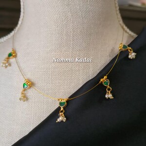 Puede incluir: Un delicado collar dorado con colgantes verdes en forma de coraz&oacute;n y peque&ntilde;os detalles de perlas blancas. El collar se exhibe en un maniqu&iacute;, con el texto "Namma Kadai" visible. El collar est&aacute; sobre una tela negra.