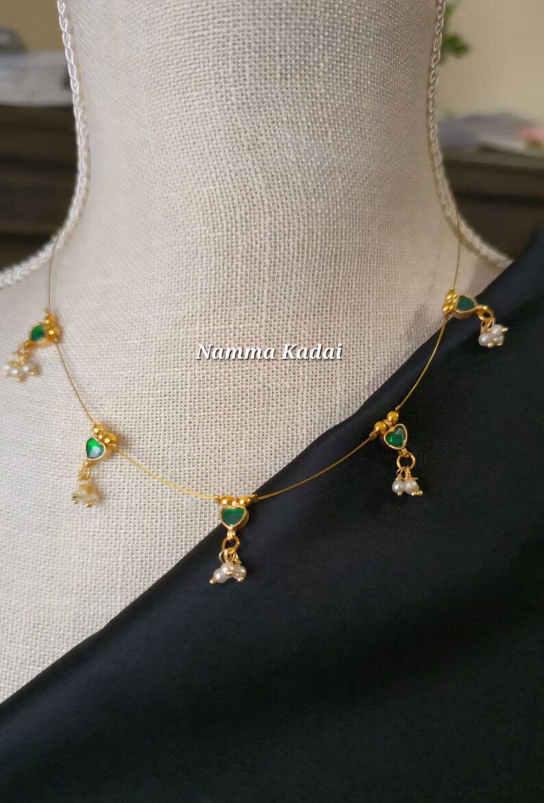 Puede incluir: Un delicado collar dorado con colgantes verdes en forma de coraz&oacute;n y peque&ntilde;os detalles de perlas blancas. El collar se exhibe en un maniqu&iacute;, con el texto "Namma Kadai" visible. El collar est&aacute; sobre una tela negra.