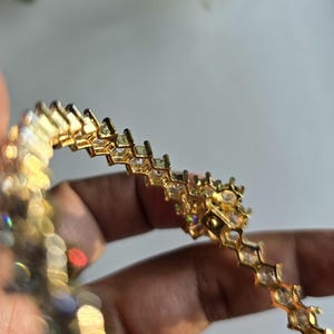 Puede incluir: Una pulsera de tono dorado que presenta una serie de eslabones geom&eacute;tricos interconectados, cada uno engastado con una piedra transparente facetada. El dise&ntilde;o muestra un patr&oacute;n angular repetitivo con un acabado met&aacute;lico que refleja la luz.
