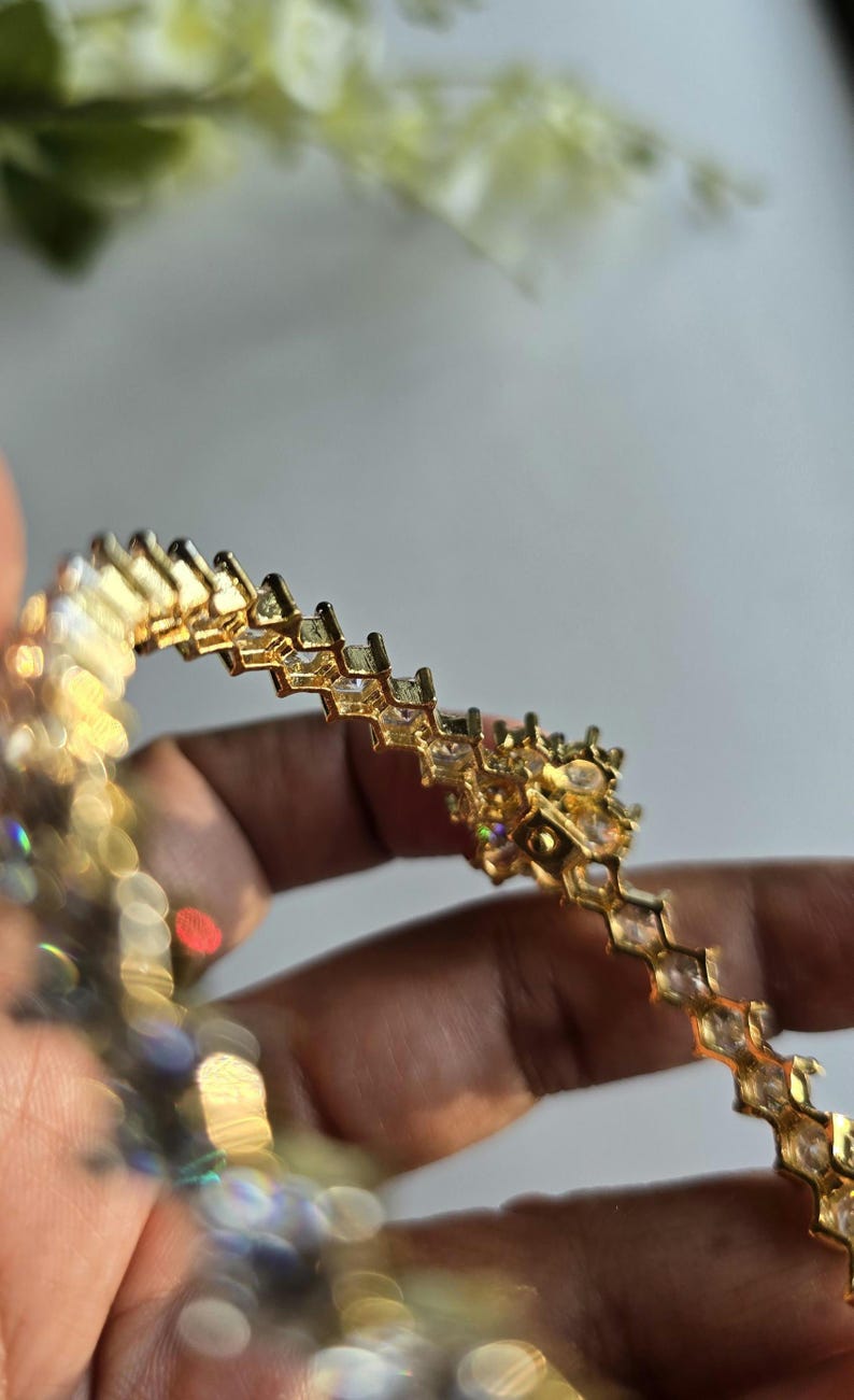 Puede incluir: Una pulsera de tono dorado que presenta una serie de eslabones geom&eacute;tricos interconectados, cada uno engastado con una piedra transparente facetada. El dise&ntilde;o muestra un patr&oacute;n angular repetitivo con un acabado met&aacute;lico que refleja la luz.