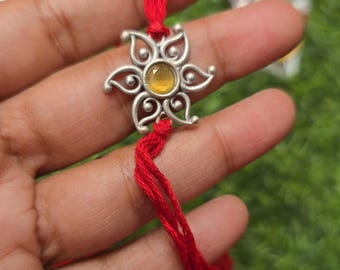 Puede incluir: Un dije floral plateado con una piedra central amarilla está unido a un hilo rojo. El dije se sostiene en una mano. El texto "Namma Kadai" es visible en la imagen.