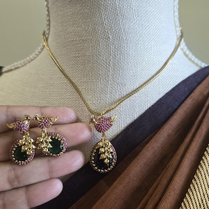 May include: A gold-toned necklace and earring set featuring a bird motif adorned with small pink stones. The pendant and earrings each include a teardrop-shaped dark green stone accented by gold-toned leaf details and a border of pink stones.