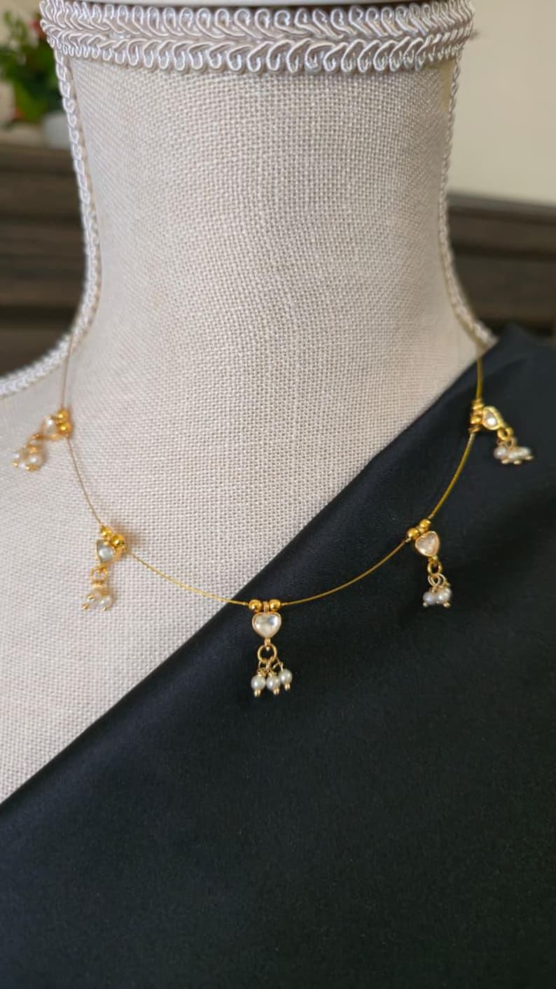 Puede incluir: Delicado collar dorado con colgantes en forma de coraz&oacute;n con piedras transparentes y peque&ntilde;os detalles de perlas. El collar se exhibe sobre un maniqu&iacute;, sobre un fondo de tela negra. Una joya sencilla y elegante.
