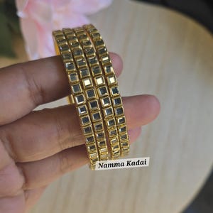 May include: Four gold-colored bangles with square-shaped embellishments are displayed. The bangles are stacked together, and the text "Namma Kadai" is visible. The jewelry is likely made of metal and is designed to be worn on the wrist.