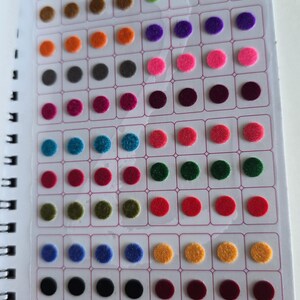 May include: A sticker sheet containing rows of small, circular, felt-like adhesive dots in a wide variety of colors including red, blue, green, yellow, orange, pink, and black. The top of the sheet features the text A QUALITY PRODUCT and STICKER KUMKUM.