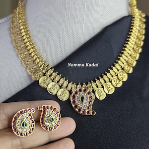 May include: Gold-toned necklace and matching earrings set. The necklace features a series of coin-like pendants and a central paisley-shaped pendant adorned with red, white, and green gemstones. The earrings mirror the paisley design. The text "Namma Kadai" is visible.