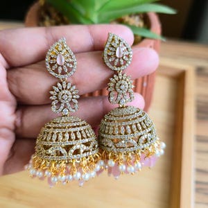 May include: Gold-toned, ornate earrings with a teardrop top, floral center, and a bell-shaped bottom. The earrings are embellished with clear and pink gemstones, and small white pearls. The earrings are held in a person's hand.