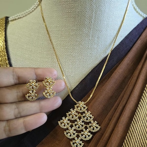 May include: A gold-toned necklace and earring set featuring a pendant and matching stud earrings. The pendant is composed of multiple interconnected fan-shaped segments encrusted with small clear stones, suspended from a thin gold-toned chain.