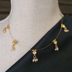 Puede incluir: Delicado collar de oro con colgantes en forma de coraz&oacute;n con piedras transparentes y peque&ntilde;os detalles de perlas. El collar se exhibe sobre un maniqu&iacute;, sobre un fondo de tela negra. Una pieza de joyer&iacute;a sencilla y elegante.