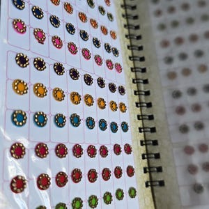 May include: A spiral-bound booklet containing rows of adhesive bindi stickers in various colors including green, orange, blue, pink, purple, yellow, and red. Each circular sticker features a metallic gold-toned scalloped border around a colored center.