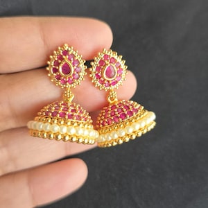 May include: Gold-toned earrings with a ruby-red gemstone design. The earrings feature a teardrop-shaped ruby at the top and a bell-shaped bottom adorned with small pearls. The earrings are held against a dark background.
