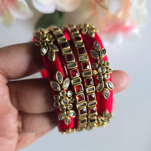 May include: A set of three vibrant red silk-wrapped bangles featuring gold-toned metal accents. The bangles are decorated with clear, leaf-shaped crystals arranged in floral patterns and square-cut red stones set in a metallic frame.