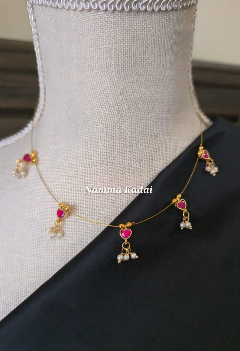 Puede incluir: Delicado collar de oro con colgantes rosas en forma de coraz&oacute;n y peque&ntilde;os grupos de perlas blancas. El collar se exhibe en un maniqu&iacute;, con el texto "Namma Kadai" visible.