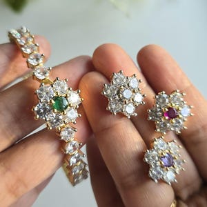 May include: A gold-toned jewelry set consisting of a bracelet and three stud earrings. Each piece features a floral design with clear white stones surrounding a central colored stone, including green, red, and purple accents in a cluster setting.