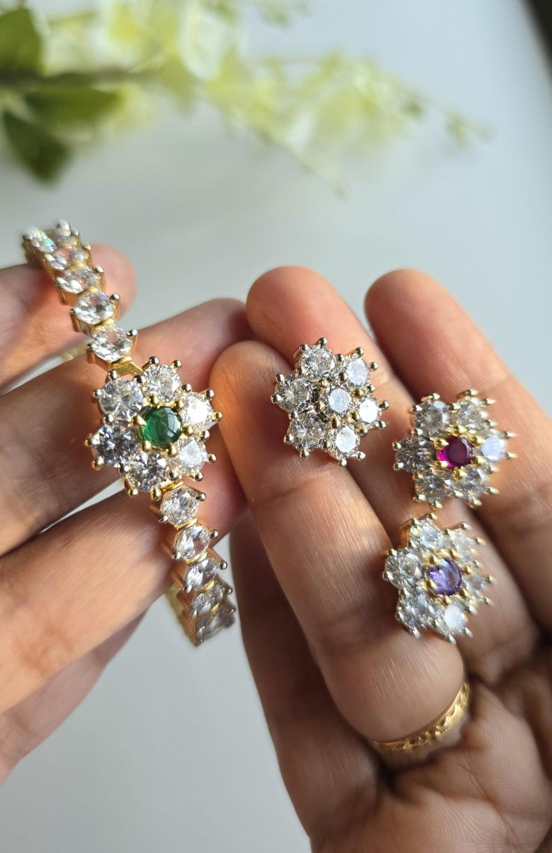 Puede incluir: Un conjunto de joyer&iacute;a en tono dorado que consta de una pulsera y tres pendientes. Cada pieza presenta un dise&ntilde;o floral con piedras blancas transparentes que rodean una piedra central de color, incluyendo acentos en verde, rojo y morado.