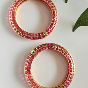 May include: Two gold bangles with a vibrant pink gemstone setting. The gemstones are square-cut and arranged closely together, creating a continuous band of color. The bangles are displayed on a white surface.