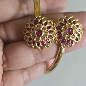 May include: A gold-toned open cuff bracelet featuring two prominent floral-shaped ornaments. Each flower is encrusted with small, clear, reflective stones and centered with a vibrant, deep pinkish-red stone, creating a symmetrical design.