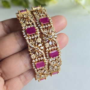 May include: Two gold-tone bangles featuring rectangular fuchsia pink stones and clear white accent stones arranged in a floral pattern. The metal surface is textured with intricate filigree detailing surrounding the embedded gemstones.