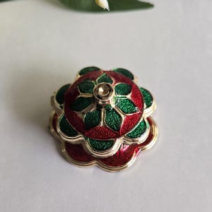 May include: A multi-layered flower-shaped metal pin with a gold-tone finish. The design features alternating sections of glittery red and green enamel, arranged in a symmetrical, stacked petal pattern with a small circular center post.