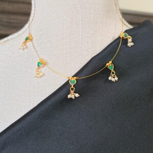 Puede incluir: Delicado collar dorado con colgantes verdes en forma de coraz&oacute;n y peque&ntilde;os detalles de perlas blancas. El collar se exhibe sobre un maniqu&iacute; con un fondo de tela negra, mostrando el dise&ntilde;o y los detalles de la joya.
