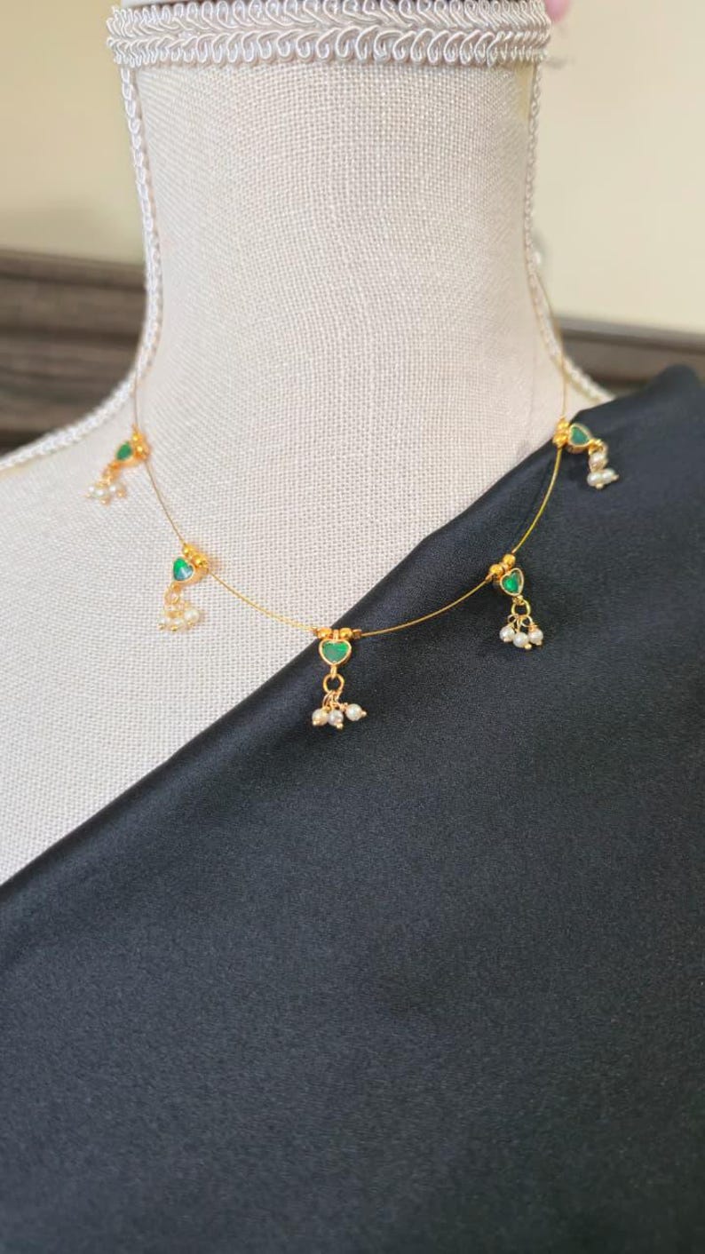 Puede incluir: Delicado collar dorado con colgantes verdes en forma de coraz&oacute;n y peque&ntilde;os detalles de perlas blancas. El collar se exhibe sobre un maniqu&iacute; con un fondo de tela negra, mostrando el dise&ntilde;o y los detalles de la joya.